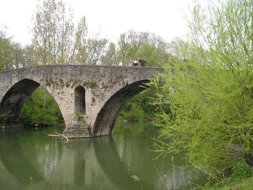 Two friendly pilgrims walking across a bridge in lush green Camino surroundings